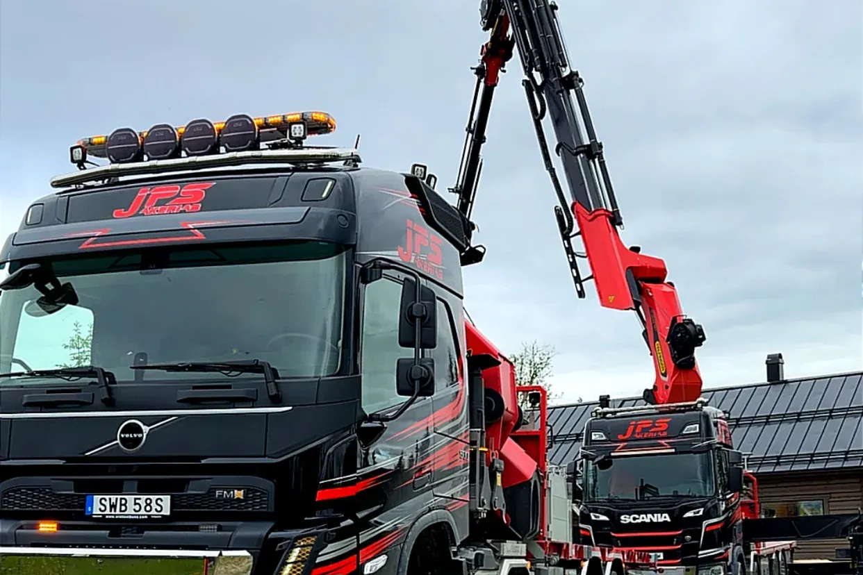 Svart JPS-lackad Volvo kranbil med utfälld röd kran, och en Scania kranbil bakom.