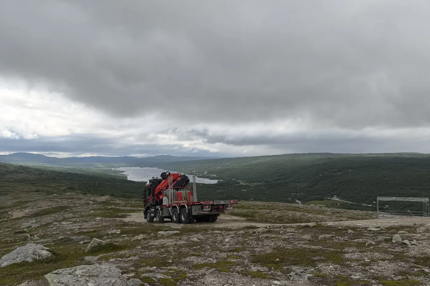 Kranbil från JPS uppställd på kalfjäll med sjö och skogklädd dalgång nedanför.